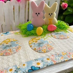 Może przedstawiać: Biały bieżnik stołowy z pikowanym wzorem przedstawiającym kwiatowe motywy i koła. Dwie pastelowe figurki królików i kolorowe pisanki wielkanocne znajdują się na tle zielonej trawy.