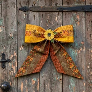 May include: A decorative fabric bow featuring a sunflower center and a pattern of autumn leaves and flowers in shades of yellow, orange, and brown. The bow has two wide loops and two long, pointed tails, with a textured finish on the fabric.