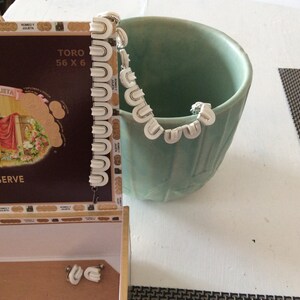 May include: A white plastic necklace with a scalloped design, resting in a green ceramic bowl. The bowl is on a white surface with a black and white patterned placemat.