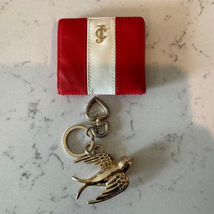 May include: A red and white striped leather wallet with a gold J.C. logo. A gold bird keychain with a heart charm is attached to the wallet.