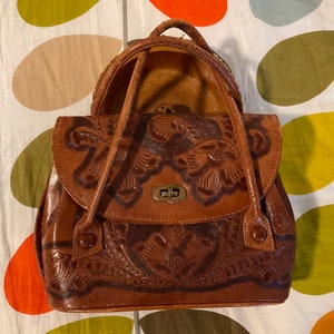 May include: A brown leather handbag with intricate floral tooling. The bag has two top handles and a flap closure with a metal clasp.