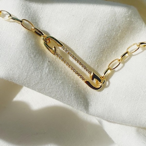 Könnte beinhalten: Goldfarbene Halskette mit einem Sicherheitsnadel-Anhänger, verziert mit kleinen, funkelnden Steinen. Die Halskette hat ein filigranes Ketten-Design und wird vor einem weißen Stoffhintergrund präsentiert. Der Sicherheitsnadel-Anhänger ist das zentrale Element.