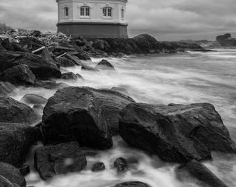 May include: Black and white photograph of a lighthouse on a rocky coastline. The lighthouse is white with a red base and a black lantern room. The ocean waves crash against the dark rocks in the foreground under a cloudy sky.