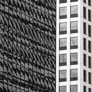 May include: Black and white architectural photograph of two adjacent buildings. One building has a reflective glass facade with a repeating pattern. The other building has a white facade with evenly spaced rectangular windows.