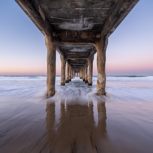 May include: A photograph of a concrete pier stretching out over the sea. The image is taken from a low angle, showing the pier's supports and the water flowing around them. The sky displays a gradient of blue and pink hues.