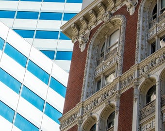May include: Architectural detail of a building with red brick and ornate white trim. The building has arched windows and decorative elements. In the background, a modern building with white panels and blue windows is visible against a clear blue sky.
