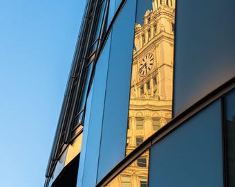 May include: A modern building with reflective glass panels, mirroring a historic clock tower. The tower's details, including the clock face and architectural features, are clearly visible in the reflection against a clear blue sky.