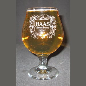 May include: A clear glass beer goblet filled with a light amber liquid. The glass has a stem and a wide bowl, with the words "HAAS Brewhaus Est. 2015" etched in white. The glass is on a dark background.