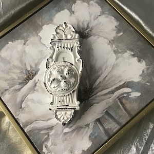 May include: White decorative wall hook with a distressed finish, mounted on a floral print canvas in a gold frame. The hook features a circular design with floral detailing and a decorative top and bottom.