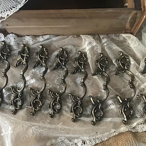 May include: A collection of eight antique bronze-toned drawer pulls with an ornate, scrolling design. The pulls have a curved handle and decorative flourishes, perfect for adding a vintage touch to furniture. They are arranged on a lace-covered surface.