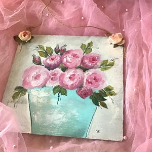 May include: A floral painting featuring pink roses in a turquoise vase. The artwork is on a rectangular wooden panel, with green leaves and rose buds. Two small, artificial roses are attached to the top corners. The background is a pink, sheer fabric.