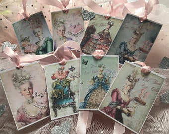 May include: A collection of rectangular gift tags featuring vintage illustrations of a woman in ornate gowns and hairstyles. Each tag has a pink ribbon and the phrase "Let Them Eat Cake". The tags are decorated with glitter and rhinestones.