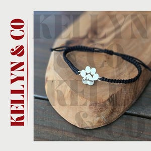 May include: A black braided bracelet with a silver paw print charm. The charm has the name "Autumn" engraved on it. The bracelet is displayed on a wooden surface.