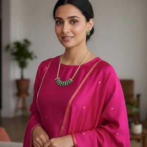 May include: A woman wearing a magenta-colored outfit, including a long-sleeved top and a matching scarf. She is wearing a gold necklace with green beads and matching green earrings. The outfit has gold trim.