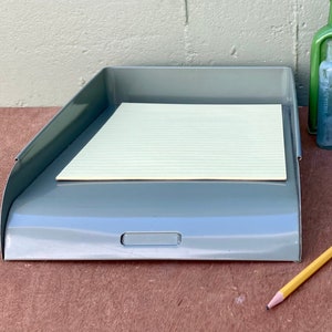 May include: A gray metal desk tray holding a single sheet of lined off-white paper. Beside the tray are three glass bottles in green, clear, and blue, along with a yellow wooden pencil with a pink eraser, all arranged on a flat surface.