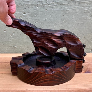 May include: A dark brown, carved wooden ashtray with a bear-shaped design. The bear is carved from a single piece of wood and has a textured surface. The ashtray has a circular depression for holding ashes and a small, round holder.