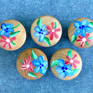 May include: Five wooden cabinet knobs with hand-painted floral designs. Each knob features a light wood grain base with blue and pink flowers and green leaves. The knobs are arranged on a blue denim surface.