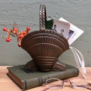 May include: A bronze-toned metal basket shaped like a fan or shell with a braided handle. It contains a small branch with red berries, a vintage black and white photograph of two people, and a white ribbon. A green book and reading glasses sit nearby.