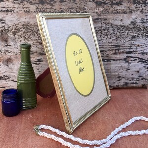 May include: A gold-colored picture frame with an oval mat, displaying the text "8 x 10 Oval Mat." Beside the frame are a green bottle, a small blue jar, and a white beaded necklace. The frame is propped up on a wooden surface.