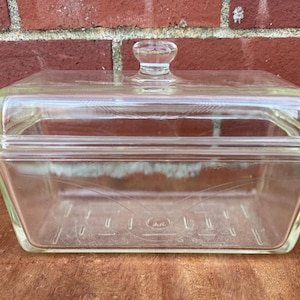 May include: A clear, rectangular glass casserole dish with a matching lid. The dish has a rounded knob on top of the lid and etched designs on the front. The dish is sitting on a wooden surface in front of a brick wall.