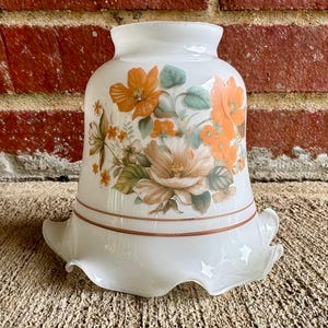 May include: White glass lampshade with a floral design featuring orange and peach flowers, green leaves, and brown accents. The lampshade has a ruffled edge and a brown band near the bottom.