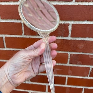 May include: A clear, vintage hand mirror with a round, beveled mirror surface and a long, ribbed handle. The mirror is held against a red brick wall, showcasing its transparent design and classic style.