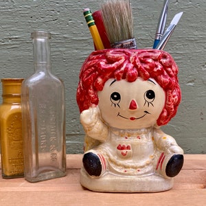 May include: A ceramic Raggedy Ann doll-shaped pencil holder with red hair and a smiling face, holding art supplies. Two antique glass bottles and a mustard-colored jar are to the left. The background is a weathered green wall.