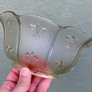 May include: A clear, textured glass bowl with a scalloped rim and a base. The bowl features embossed fleur-de-lis designs. The bowl is approximately 6 inches long and 3 inches wide.