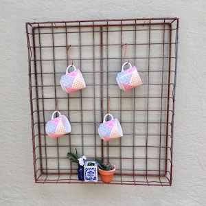 May include: A rustic, square, metal wall grid with four small, patterned teacups hanging from copper hooks. Below, a small shelf holds a blue vase, a patterned ceramic box, and a terracotta pot with a plant. The cups have a patchwork design.