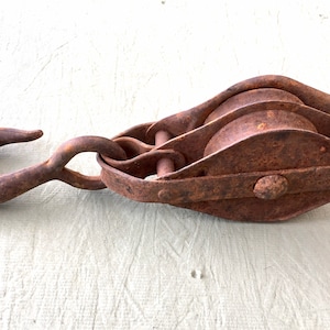 May include: A rusty, antique pulley system with a large hook. The metal components show significant wear and corrosion, indicating age and use. The pulley has multiple wheels and is designed for lifting heavy objects. The overall color is a weathered brown.