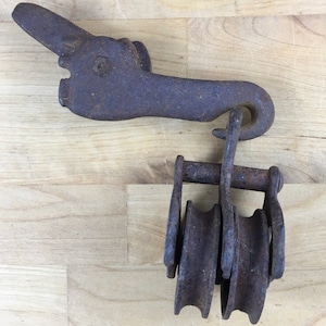 May include: A vintage, rusty metal pulley system with a hook and three wheels. The pulley has a weathered appearance, suggesting age and use. The background is a light-colored wooden surface.