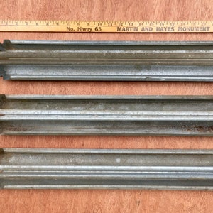 May include: Three weathered, corrugated metal pieces with a ridged design, possibly for a construction or industrial application. Each piece has a tag attached. A wooden ruler with text is in the background.