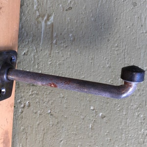 May include: A metal coat hook with a square mounting plate and a curved arm. The hook is attached to a wooden surface and has a black, conical end. The metal shows signs of rust.