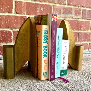May include: Two gold-colored metal bookends with a pointed, triangular silhouette holding four books titled The Children's Busy Book, Devotions from the Garden, Traditional Tales of Long, Long Ago, and 1,001 Symbols between them.