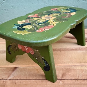 May include: A small, green wooden footstool with a floral design. The top is painted with colorful flowers and leaves in shades of yellow, pink, blue, and green. The legs are also green and feature painted floral accents.