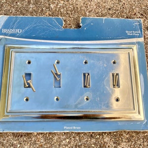 May include: A plated brass quad switch wall plate with a rectangular design. The plate has four switch openings and includes screws for installation. The packaging is blue with the Brainerd brand name.