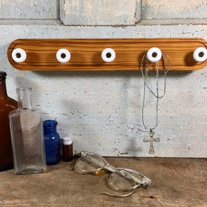 May include: A wooden wall-mounted coat rack with five white ceramic knobs. A silver necklace with a cross hangs from one knob. Various antique bottles and eyeglasses are arranged below the rack.