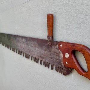 May include: A long, vintage crosscut saw featuring a weathered, dark metal blade with prominent, sharp teeth. The tool includes a polished wooden handle at one end and a secondary, vertical wooden handle attached to the top of the blade.