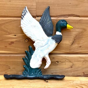 May include: A cast iron duck sculpture with outstretched wings, painted in white, black, and teal. The duck has a yellow beak and a green head. The sculpture is mounted on a black base and is a decorative item.