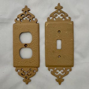 May include: Two antique, ornate light switch covers. One cover has two circular openings for an electrical outlet, and the other has a rectangular opening for a light switch. Both are a textured, tan color with decorative scrollwork.