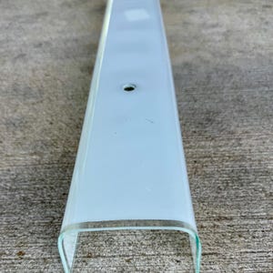 May include: A long, rectangular light fixture cover made of frosted white glass with a curved, U-shaped profile. The glass features a single, centered circular hole for mounting purposes, with clear, transparent edges along the bottom sides.