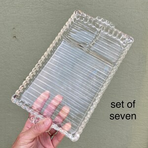 May include: A rectangular clear glass serving tray featuring a ribbed texture on the surface and a scalloped edge design. The tray is divided into two sections, one larger and one smaller. The text "set of seven" is printed on the right side.