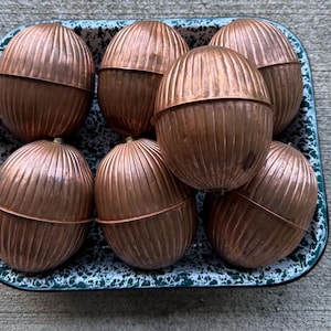 May include: Seven copper-colored, ribbed, oval-shaped containers with horizontal seams, arranged in a rectangular, white and green speckled enamel tray. Each container features a rounded top and bottom section, creating a uniform, metallic appearance.