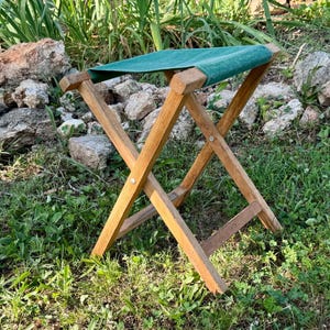 May include: A folding stool with a natural wood frame and a dark green fabric seat. The stool features a cross-leg design with metal hardware connecting the wooden supports, creating a portable seating structure with a rectangular top surface.