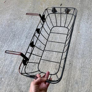 May include: A black metal bicycle basket with a rectangular shape and curved edges. The basket has a grid-like design and features two red-brown metal arms for mounting. The basket is held by a hand.