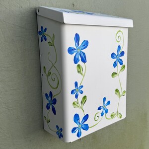 May include: A white metal wall-mounted mailbox featuring hand-painted blue flowers with green stems and swirling vines. The rectangular box has a hinged lid at the top and a clean, smooth finish, displaying a floral motif on the front and top.