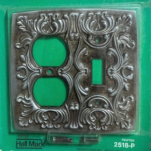 May include: A pewter-colored, ornate light switch cover with two electrical outlets and a single light switch. The cover features a detailed, scrolling floral design. The background is green.