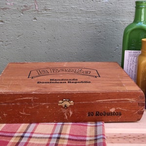 May include: A wooden cigar box with the text "La Bamba Handmade Dominican Republic" and "20 Robustos". Beside it are three glass bottles in green, yellow, and blue, and a vintage postcard.