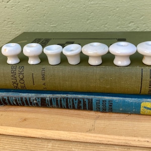 May include: A collection of eight white ceramic knobs with a mushroom-like design, arranged on top of two vintage books. The books are green and blue, with visible text. The knobs are likely for furniture or cabinetry.