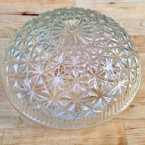 May include: A clear, glass light fixture cover with a textured, starburst pattern. The cover is dome-shaped and sits on a light-colored wooden surface. The glass has a decorative, geometric design.